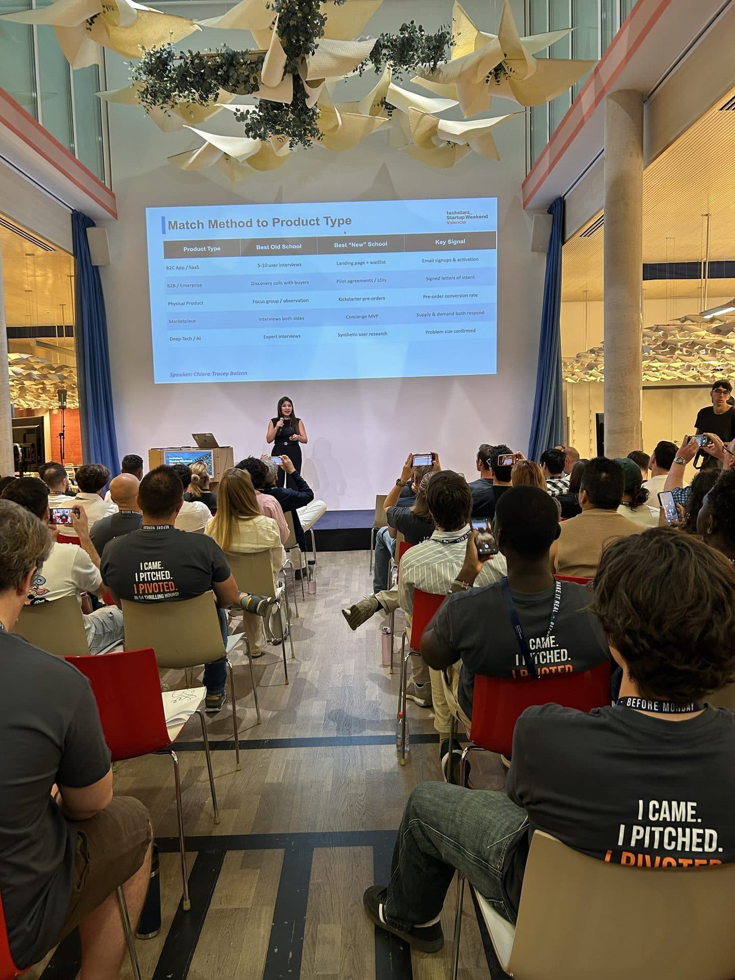 A speaker presents on stage beside a projector screen to a seated audience, June 2026.